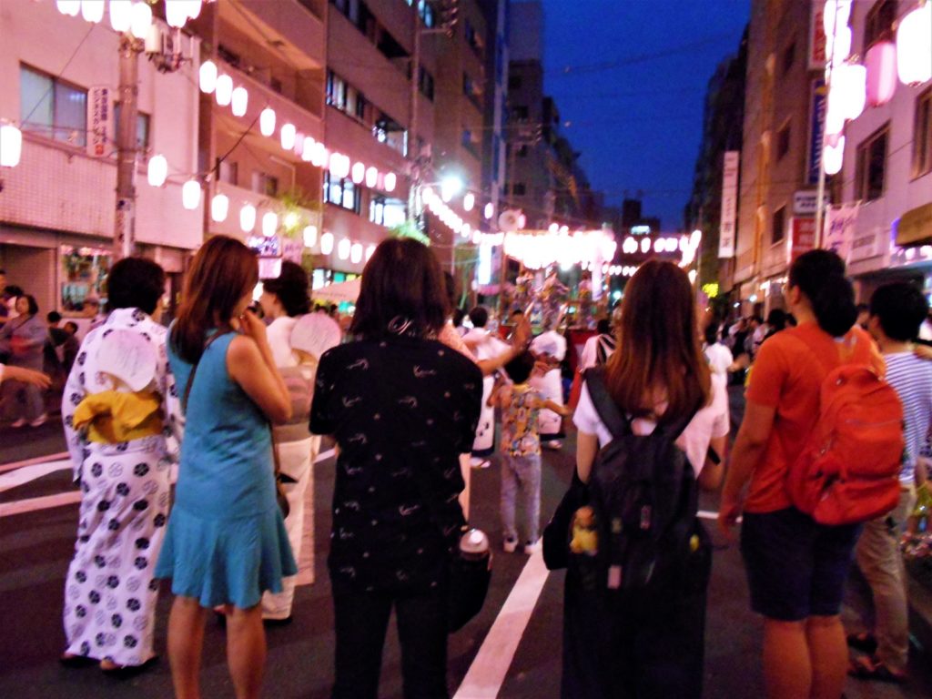 盆踊りする人たちと、それを眺める外国人旅行客の女性4人。
